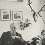 Johann Dietrich Lauenstein sitzt an seinem Schreibtisch mit Telefon, im Hintergrund Gemälde und ein Hirschgeweih.