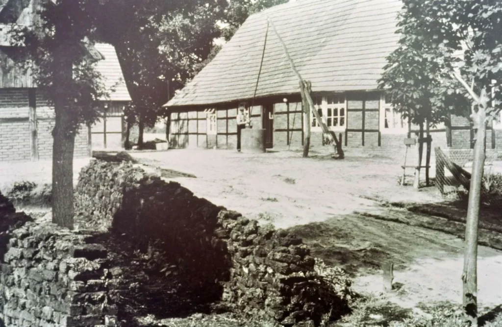 Schwarzweißfoto eines Fachwerkgehöfts mit Sandweg und Trockensteinmauer im ländlichen Emsland um 1950.