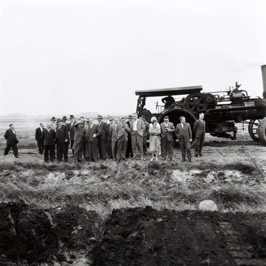 Gruppe von Männern in Anzügen besichtigt einen Dampfpflug auf moorigem Feld im Emsland, Mai 1961.