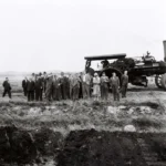 Gruppe von Männern in Anzügen besichtigt einen Dampfpflug auf moorigem Feld im Emsland, Mai 1961.