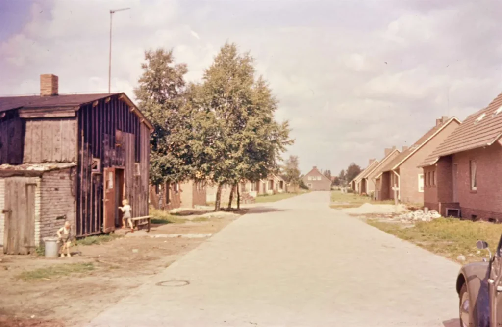 Straße in Füchtenfeld mit altem Holzschuppen links, neuen Backsteinreihenhäusern rechts und spielenden Kindern, um 1961.