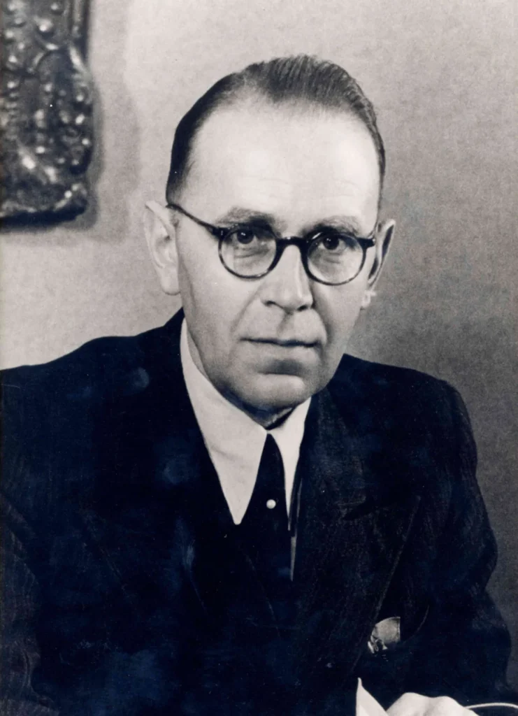 Schwarzweißes Porträtfoto eines älteren Mannes mit runder Brille und dunklem Anzug aus der Nachkriegszeit.