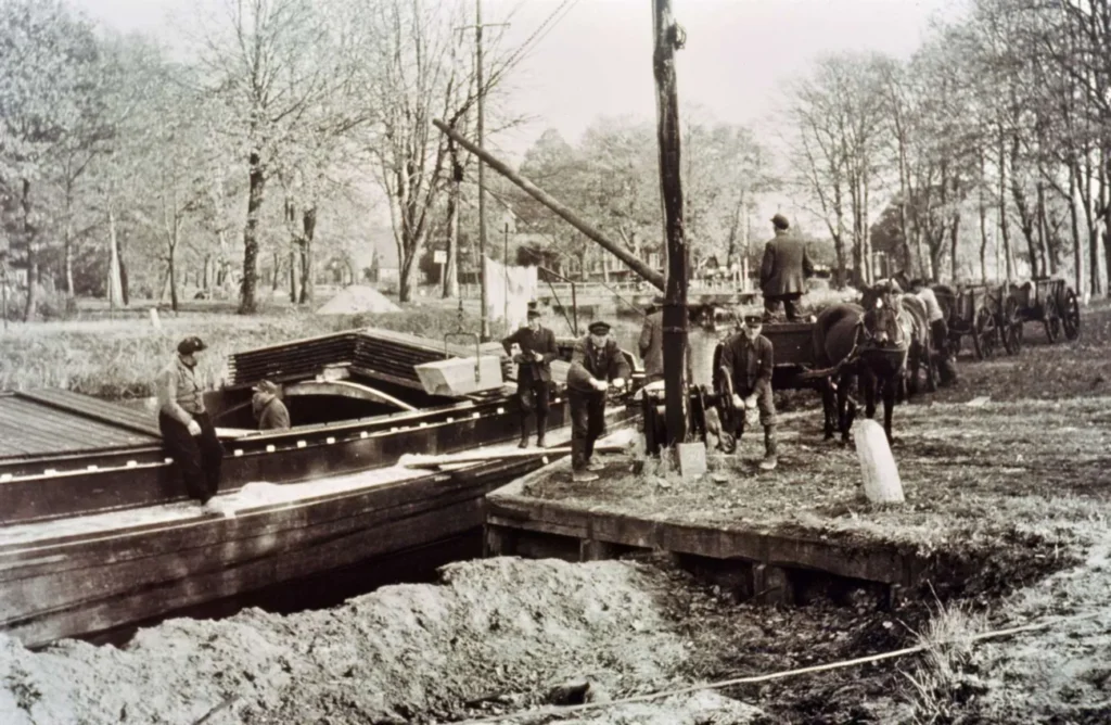 Historische Schwarzweißaufnahme von Arbeitern beim Kanalbau mit Pferdefuhrwerken und Holzplanken im Emsland.