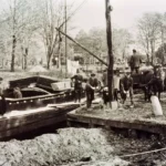 Historische Schwarzweißaufnahme von Arbeitern beim Kanalbau mit Pferdefuhrwerken und Holzplanken im Emsland.