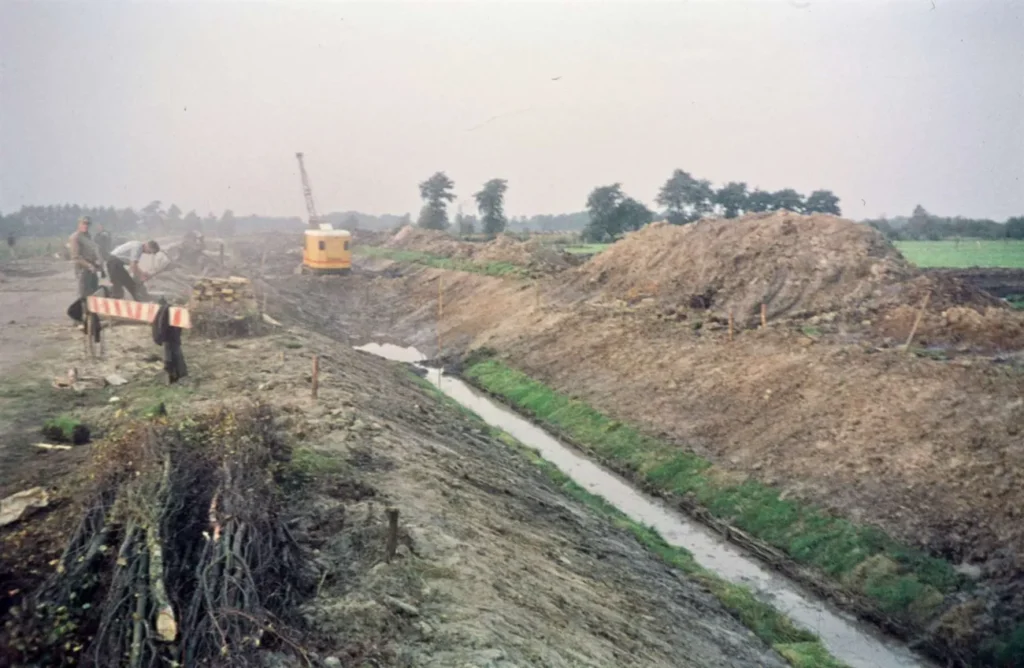 Arbeiter beim Ausheben eines Entwässerungsgrabens im Moorgebiet, mit Bagger und aufgeschütteten Erdwällen im Hintergrund.
