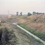 Arbeiter beim Ausheben eines Entwässerungsgrabens im Moorgebiet, mit Bagger und aufgeschütteten Erdwällen im Hintergrund.