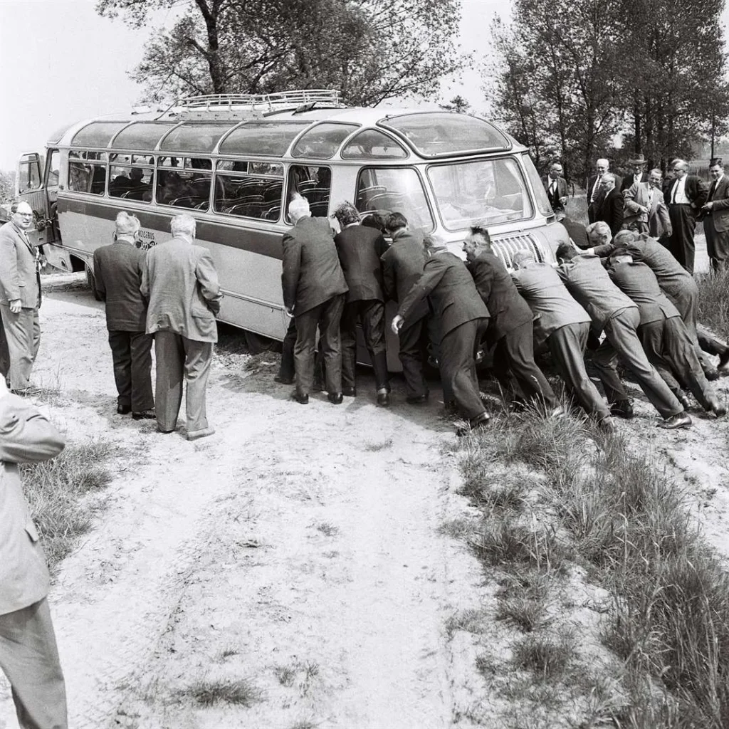 Zahlreiche Männer in Anzügen schieben gemeinsam einen im Sand steckengebliebenen Reisebus, Grafschaft Bentheim 1961.