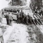 Zahlreiche Männer in Anzügen schieben gemeinsam einen im Sand steckengebliebenen Reisebus, Grafschaft Bentheim 1961.