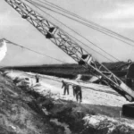 Ein Dragline-Bagger hebt Torfmassen im Emsland aus, während Arbeiter die Entwässerungsarbeiten begleiten.