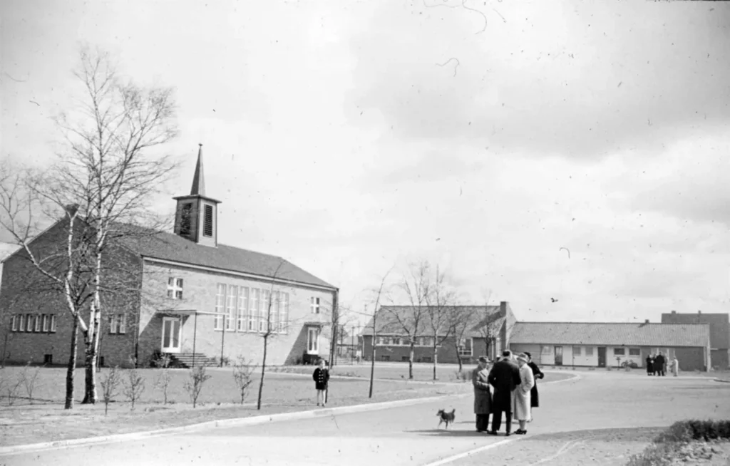 Schwarzweißfoto einer Backsteinkirche mit Spitzturm in Neugnadenfeld, davor Dorfplatz mit Bäumen und Passanten.
