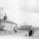 Schwarzweißfoto einer Backsteinkirche mit Spitzturm in Neugnadenfeld, davor Dorfplatz mit Bäumen und Passanten.