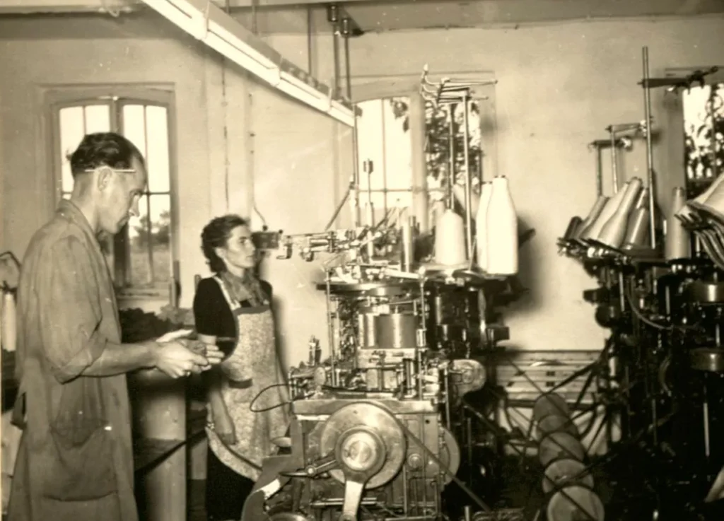 Ein Mann und eine Frau arbeiten in einer Fabrikhalle an historischen Strickmaschinen mit Garnrollen, ca. 1950er Jahre.
