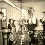 Ein Mann und eine Frau arbeiten in einer Fabrikhalle an historischen Strickmaschinen mit Garnrollen, ca. 1950er Jahre.