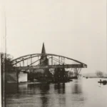 Historische Bogenbrücke über die Ems bei Hochwasser, im Hintergrund ein Kirchturm, ein Ruderboot rechts.
