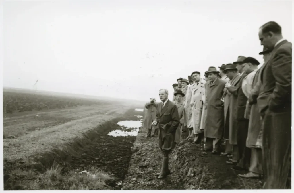Gruppe von Männern in Mänteln besichtigt ein entwässertes Emslandmoor, ein Mann zeigt in die Ferne.