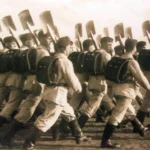Historische Schwarzweißfoto einer marschierenden Kolonne uniformierter Männer mit Spaten und Rucksäcken bei einem Aufmarsch.