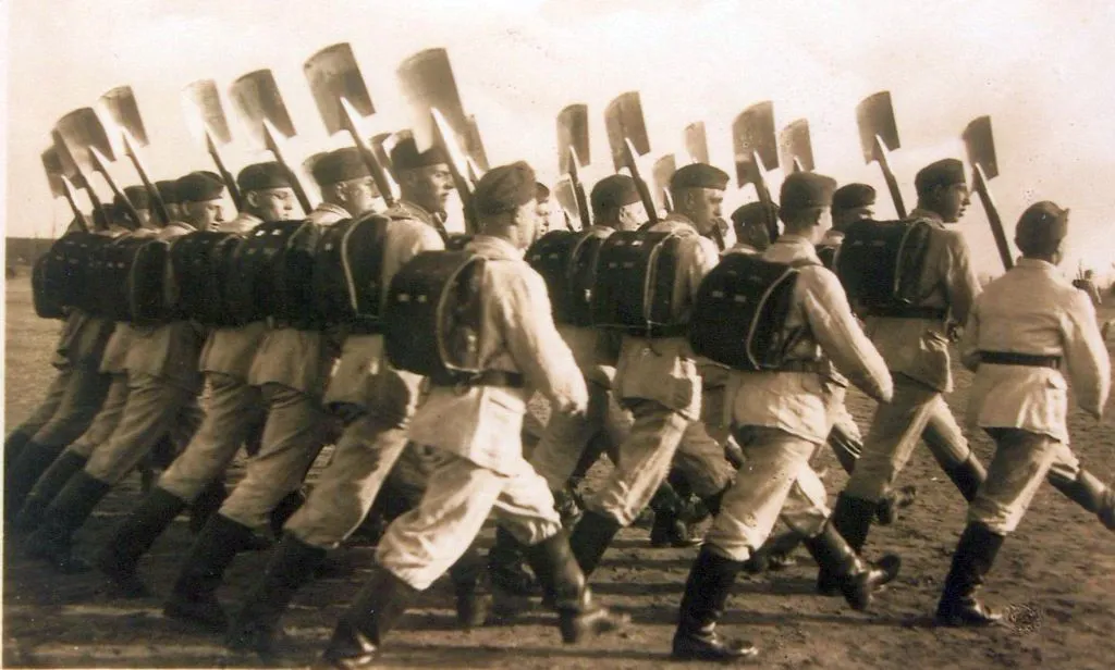 Historische Schwarzweißfoto einer marschierenden Kolonne uniformierter Männer mit Spaten und Rucksäcken bei einem Aufmarsch.