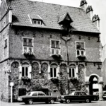 Mehrstöckiges historisches Backsteingebäude mit Bogenfenstern und Blumenschmuck, davor geparkte Autos der 1960er Jahre.