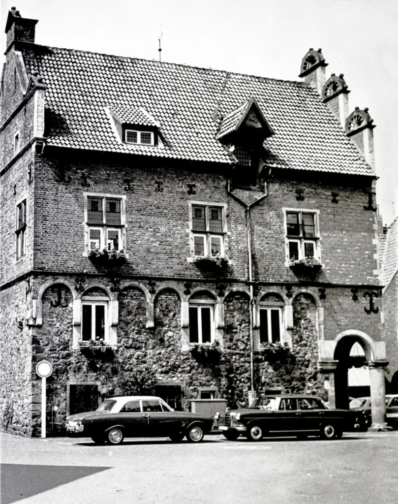 Mehrstöckiges historisches Backsteingebäude mit Bogenfenstern und Blumenschmuck, davor geparkte Autos der 1960er Jahre.