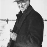 Älterer Mann mit Filzhut und Mantel steht lächelnd in winterlicher Landschaft, hält Gegenstand in den Händen.