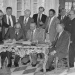 Mehrere Männer in Anzügen bei einer offiziellen Urkundunterzeichnung an einem verzierten Tisch in einem eleganten Saal.