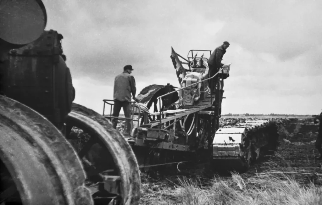 Zwei Arbeiter bedienen einen großen Kettenpflug bei der Moorkultivierung auf einem flachen Moorfeld.