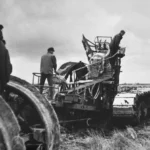 Zwei Arbeiter bedienen einen großen Kettenpflug bei der Moorkultivierung auf einem flachen Moorfeld.