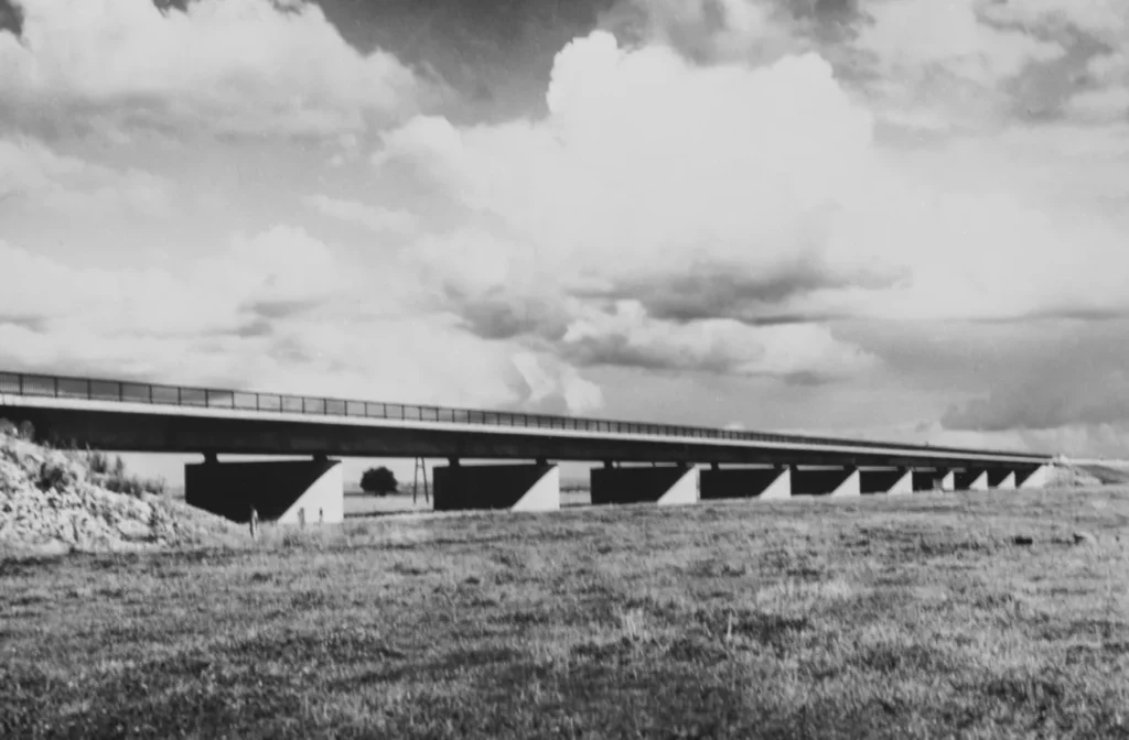 Schwarz-Weiß-Foto einer langen Betonbrücke auf Pfeilern, die über eine flache, grasige Emslandschaft führt.