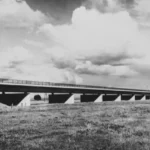 Schwarz-Weiß-Foto einer langen Betonbrücke auf Pfeilern, die über eine flache, grasige Emslandschaft führt.