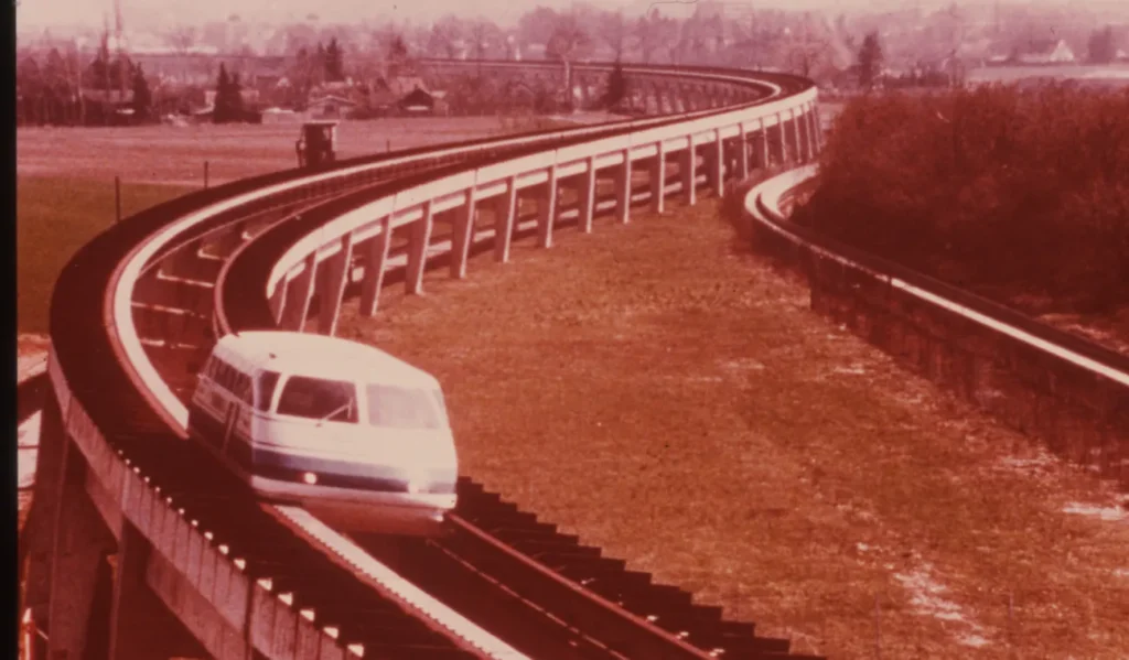 Ein weißer Transrapid-Prototyp fährt auf einer geschwungenen, erhöhten Magnetschwebebahn-Teststrecke durch flaches Land.