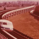 Ein weißer Transrapid-Prototyp fährt auf einer geschwungenen, erhöhten Magnetschwebebahn-Teststrecke durch flaches Land.