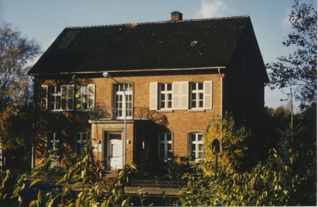 Zweigeschossige Backsteinvilla mit schwarzem Satteldach, Fensterläden und Balkon, umgeben von herbstlichen Büschen und Bäumen.