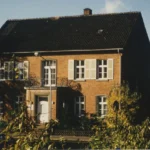 Zweigeschossige Backsteinvilla mit schwarzem Satteldach, Fensterläden und Balkon, umgeben von herbstlichen Büschen und Bäumen.