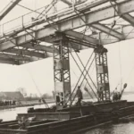 Arbeiter auf einem Ponton montieren einen Stahlträger unter einer Brückenkonstruktion über der Ems.