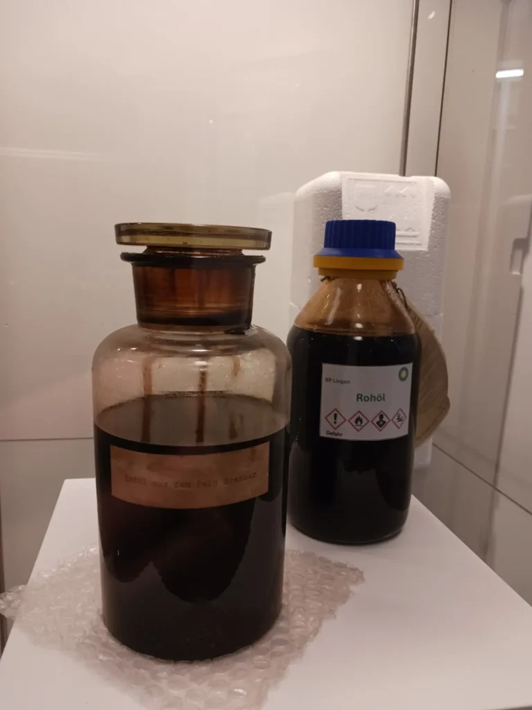 Zwei dunkle Glasflaschen mit Rohöl: eine historische Flasche mit Etikett 'Erdöl aus dem Feld Brashar' und eine moderne BP-Flasche mit der Aufschrift 'Rohöl'.