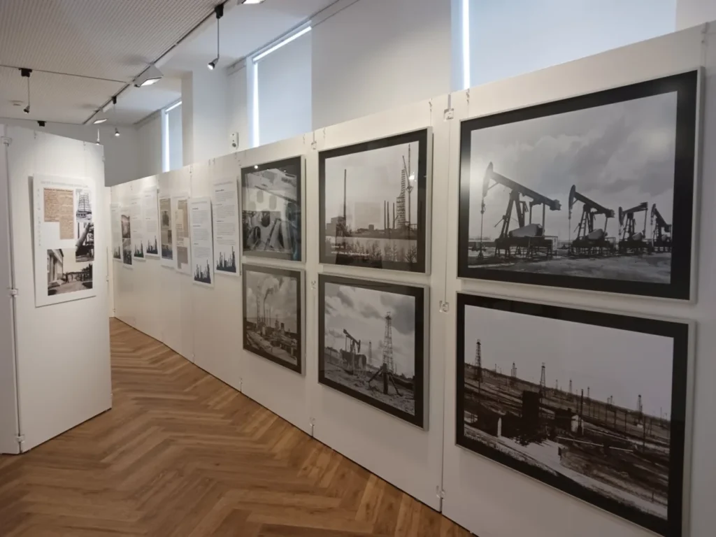 Museumsgang mit historischen Schwarzweißfotos von Ölförderanlagen und Pumpen zur Geschichte der Lingener Raffinerie.