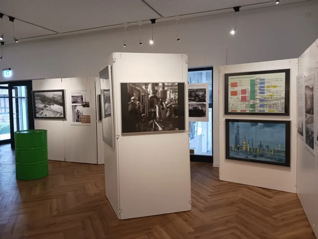 Museumsausstellung mit historischen Schwarz-Weiß-Fotos von Arbeitern, Industriediagrammen und einem grünen Ölfass.