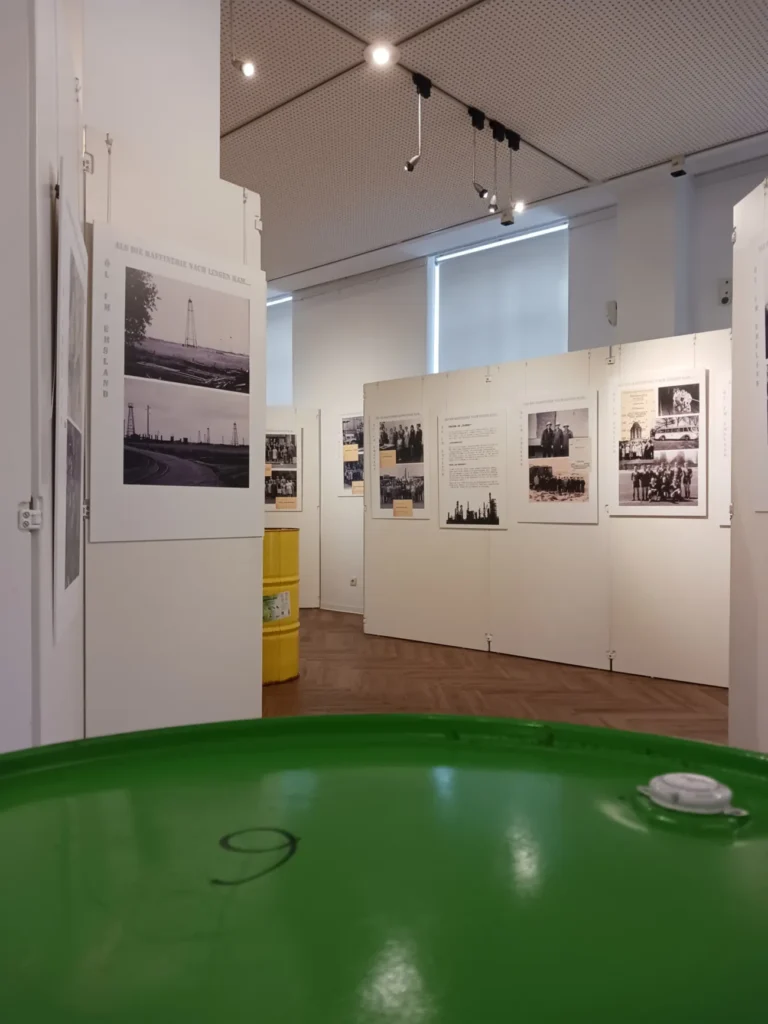 Museumsraum mit historischen Schwarz-Weiß-Fotos zur Raffineriegeschichte Lingens, im Vordergrund grünes Ölfass.