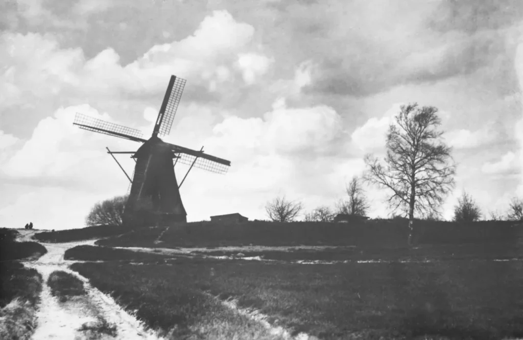 Schwarzweißfoto einer Windmühle auf einem Hügel im Emsland mit Feldweg, kahlen Bäumen und bewölktem Himmel.
