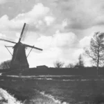 Schwarzweißfoto einer Windmühle auf einem Hügel im Emsland mit Feldweg, kahlen Bäumen und bewölktem Himmel.