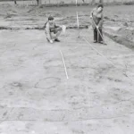 Zwei Personen dokumentieren kreisförmige Strukturen im Sand einer archäologischen Ausgrabungsstätte mit Messlatte und Schnur.