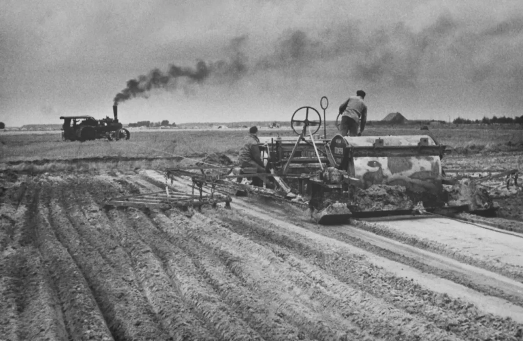 Historische Schwarzweißaufnahme: Arbeiter bedienen schwere Landmaschinen beim Umbrechen weiter Moorfelder, im Hintergrund Dampftraktor.