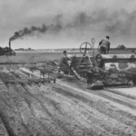 Historische Schwarzweißaufnahme: Arbeiter bedienen schwere Landmaschinen beim Umbrechen weiter Moorfelder, im Hintergrund Dampftraktor.