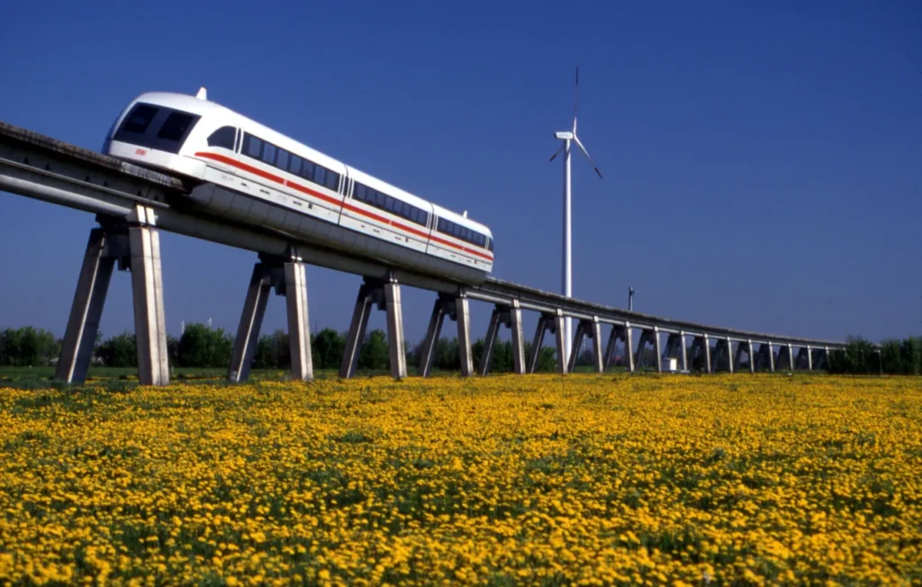 Ein weißer Transrapid-Zug gleitet auf einer Hochtrasse über ein gelbes Blumenfeld, im Hintergrund ein Windrad.