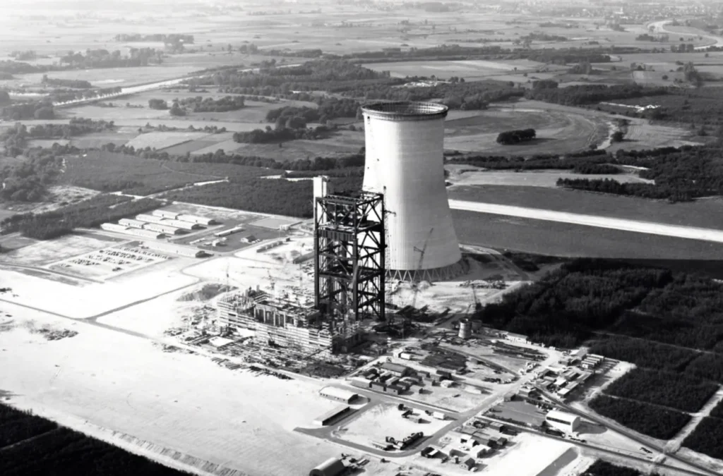 Schwarz-Weiß-Luftaufnahme eines im Bau befindlichen Industriekomplexes mit Kühlturm in flacher Landschaft.