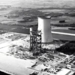 Schwarz-Weiß-Luftaufnahme eines im Bau befindlichen Industriekomplexes mit Kühlturm in flacher Landschaft.