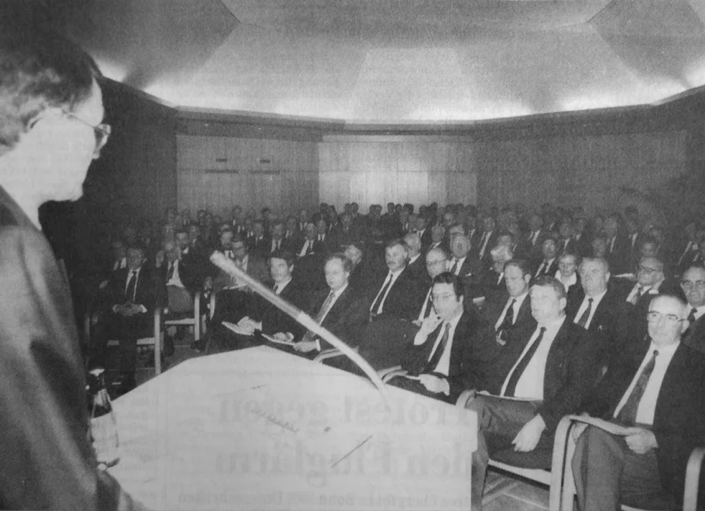 Ein Redner spricht vor einem vollbesetzten Saal mit Anzugträgern bei der Abschlussfeier der Emsland GmbH.