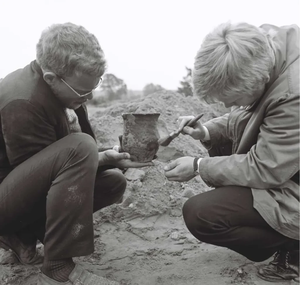 Zwei Archäologen hocken im Sand und untersuchen ein geborgenes Tongefäß mit einem Pinsel.