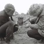 Zwei Archäologen hocken im Sand und untersuchen ein geborgenes Tongefäß mit einem Pinsel.