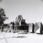 Zwei Arbeiter stehen neben einer Baumaschine auf Schienen, umgeben von Baumaterial und Kipploren auf einer Baustelle.